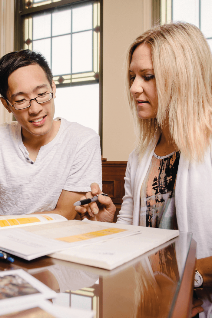 A student sits at a desk and looks at a book with an advisor. 