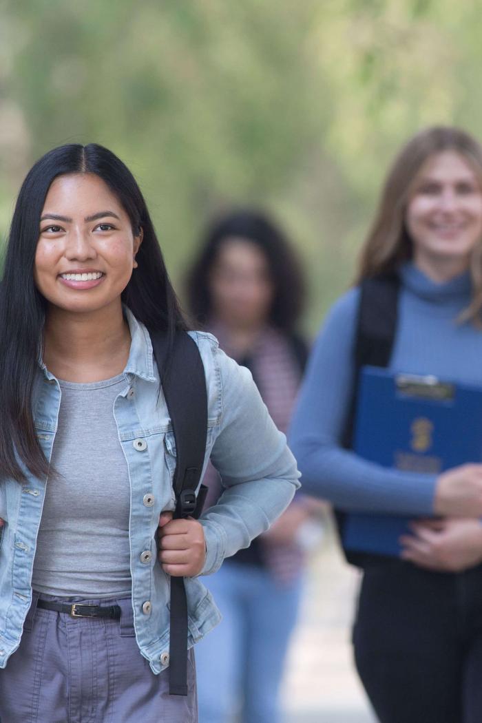 A student smiles and walks along a treed sidewalk.