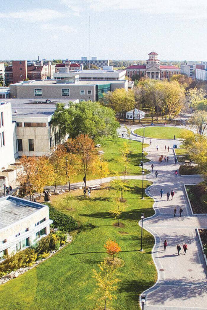 Students walk along Curry Place walkway past trees with leaves on the Fort Garry campus.