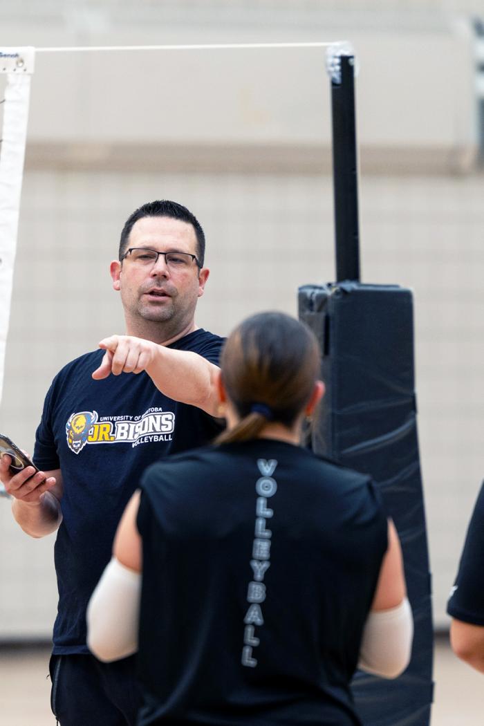 A coach points with one hand while talking to a group of young athletes circled around him.