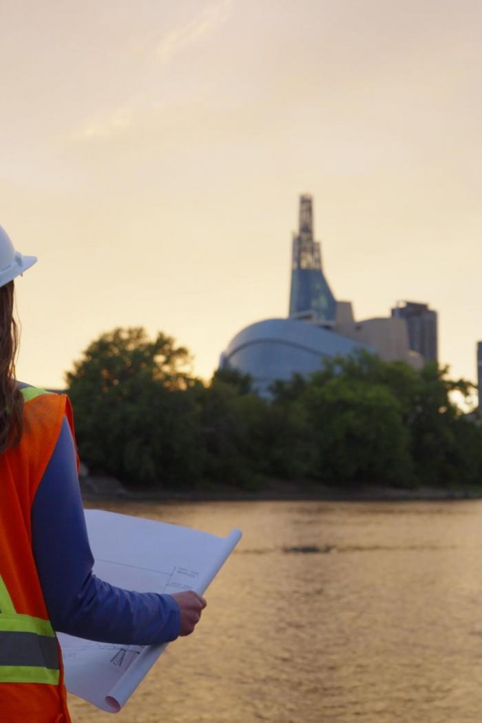 Two people with hardhats hold a technical drawing and look at the Winnipeg skyline.