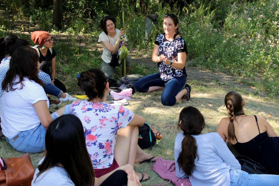 Emily McKinnon sitting on the ground outside speaking to Access Students who are sitting in a semi-circle around her.