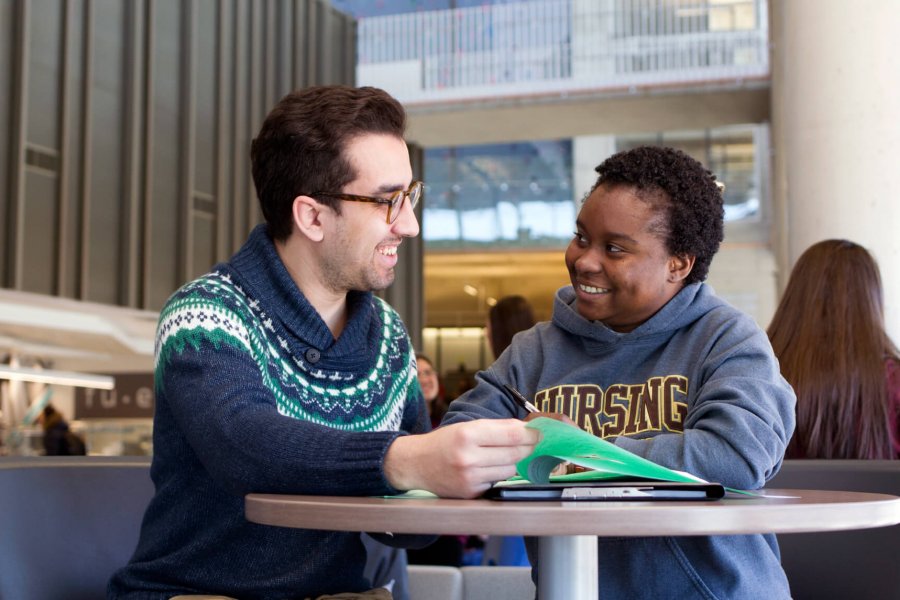 Two international students sit at a table, chatting and reviewing printed material.  