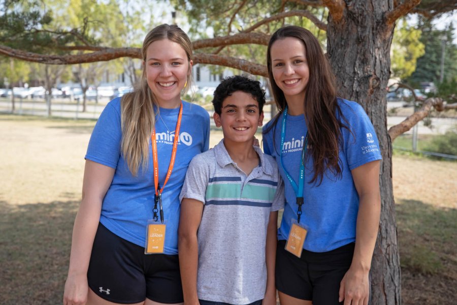 Two leaders pose for a photo with a young Mini U child outside under a tree.