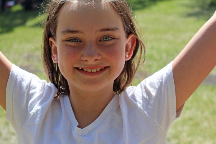 A young girl raises her arms and smiles at the camera while outdoors in a park.