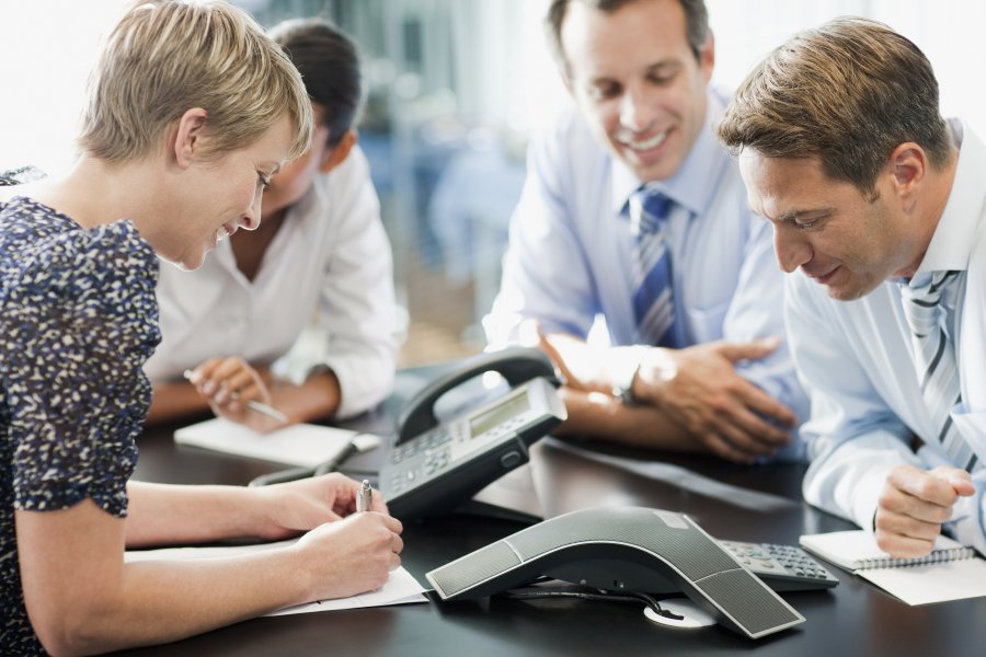 Several people in an office setting leaning close to a phone conference receiver and smiling.