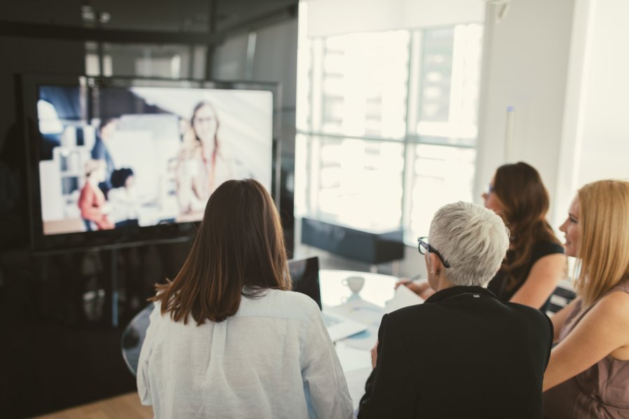 Image of several women at a table looking at a web display monitor with a woman shown