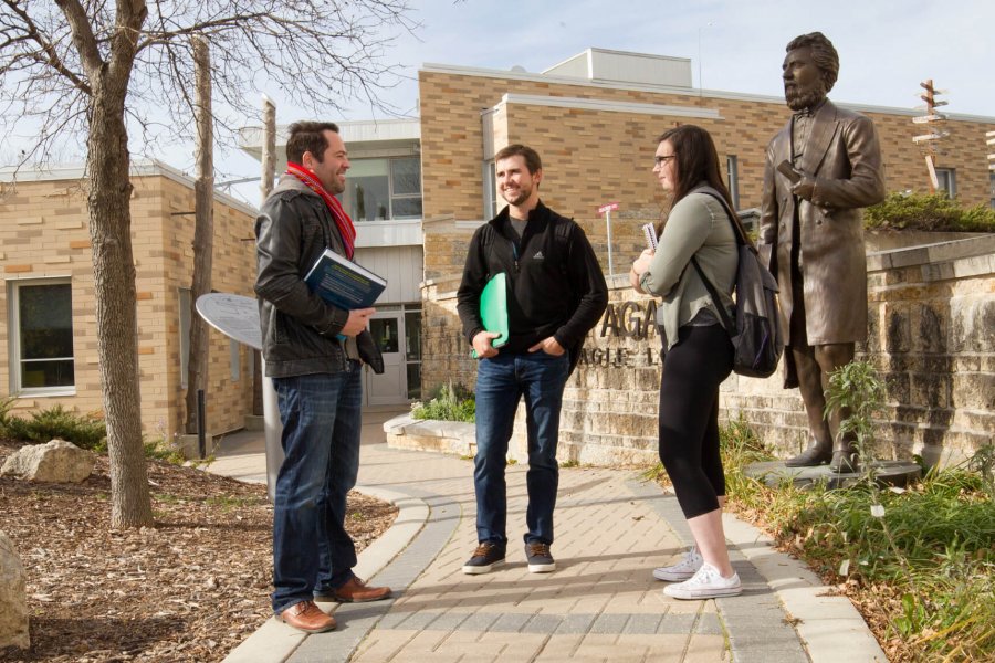 Three Indigenous students stand together talking outdoors in front of Migizii Agamik - Bald Eagle Lodge.