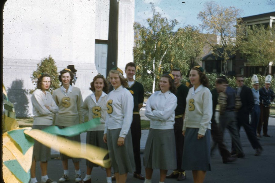 A slide of Helen Magnuson and other University of Manitoba science students in a freshie parade. Photograph shows a group of unidentified people, those in the foreground wearing matching beanie hats. Girls in the foreground appear to be wearing matching sweaters and skirts, and some of the sweaters are marked with a large "S". Image is not dated, printed on a Kodachrome slide.