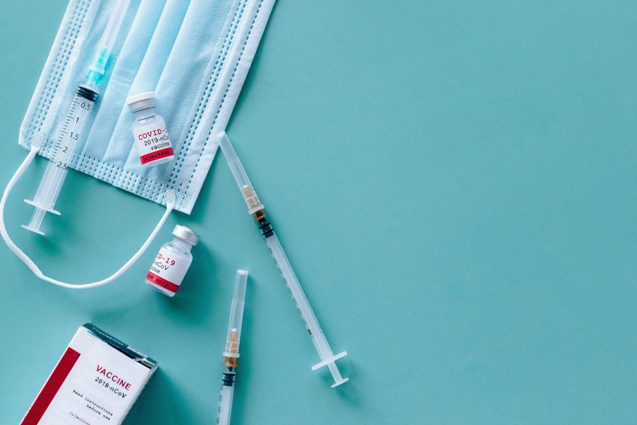 COVID-19 vaccine vials, syringes and a blue mask on a green table. Image from Pexels.