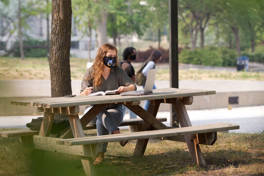 A student sits at a picnic table wearing a mask with University of Manitoba logo. Books are open on the table.
