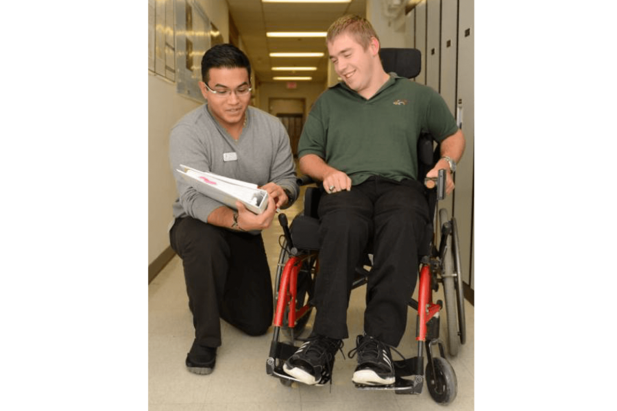 A student kneels down on one knee as he speaks to a person who is using a wheelchair and points to papers he is holding. 