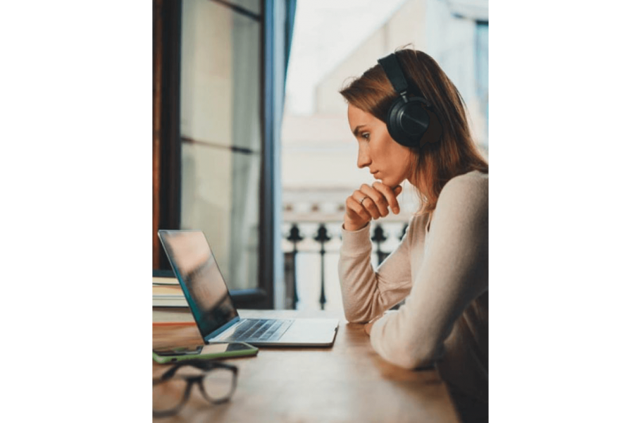 A woman wearing headphones seated at a table, leans on her elbows as she watches her laptop monitor.