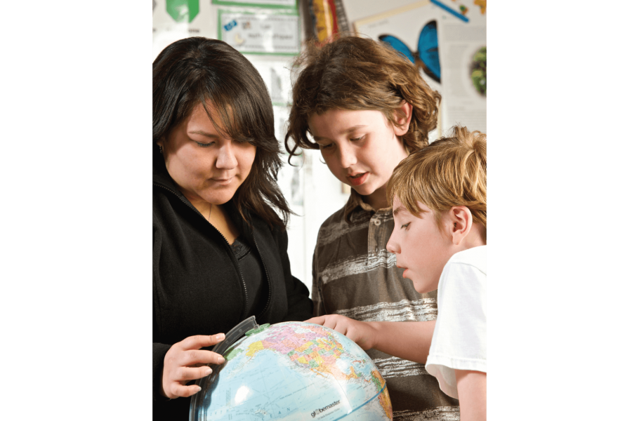 A teacher shows two young students a globe, one student points at a location.