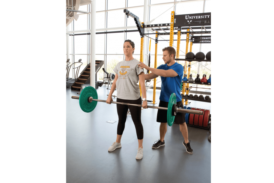 A student helps to correct the posture of a person who is lifting weights on a bar inside the Active Living Centre. 