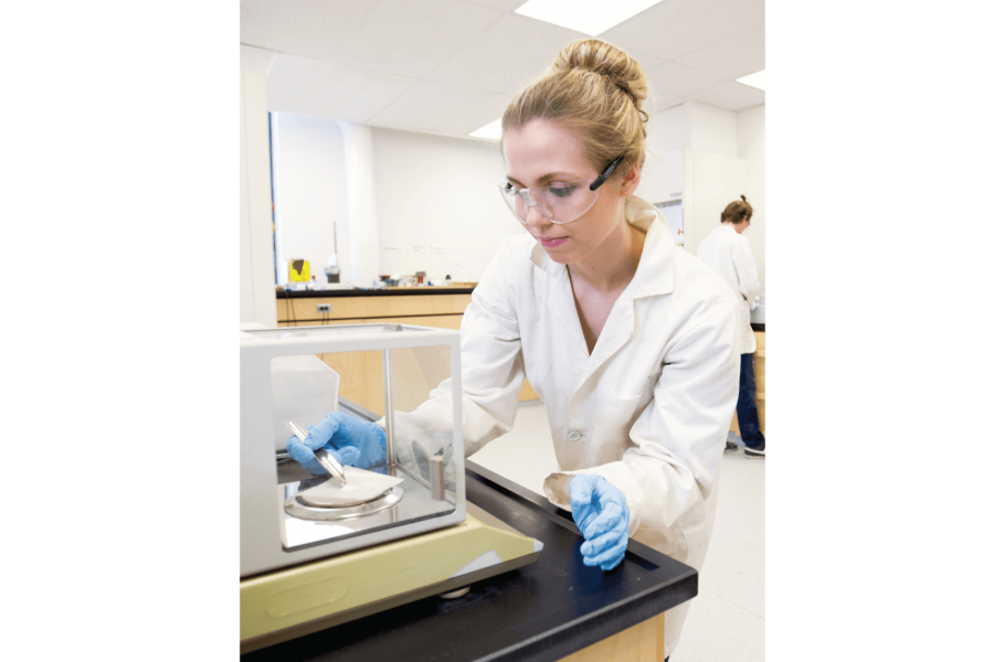 A student wearing gloves, glasses and a white coat works carefully with equipment in a lab.