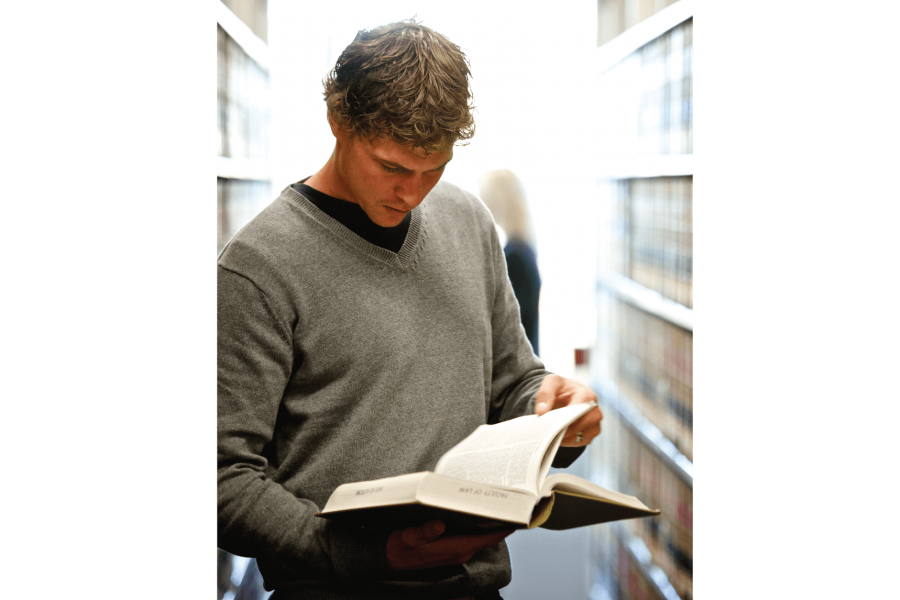 A student reads a book while standing at a bookshelf in a library.