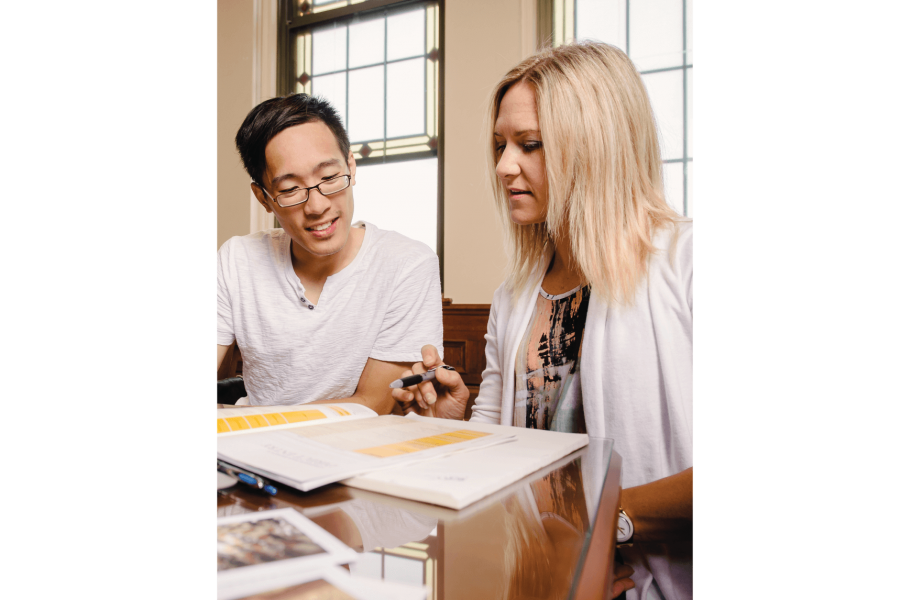 A student sits at a desk and looks at a book with an advisor. 