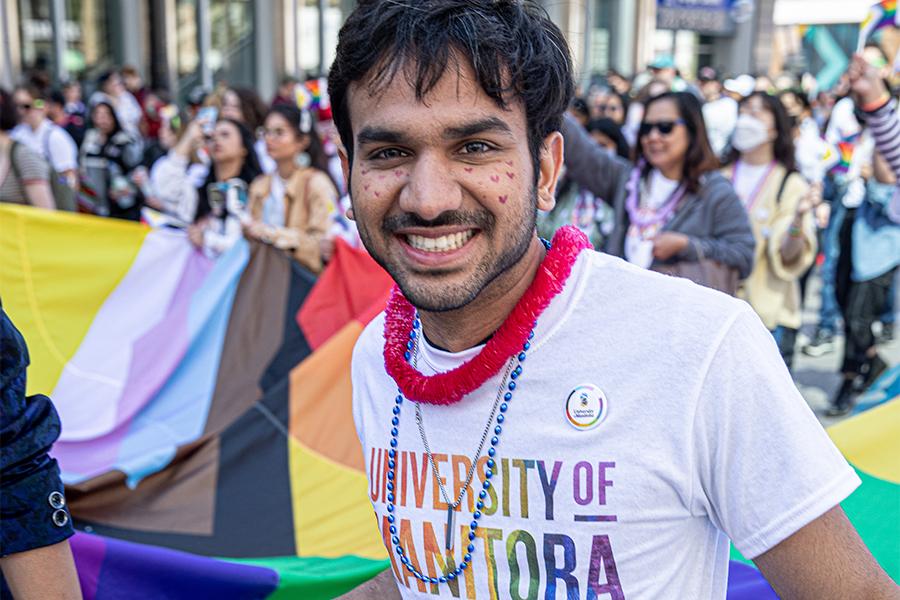 A person smiles wearing a U M rainbow shirt, a lei, and hearts drawn on their face. A very large pride flag and group of marchers are in the background.