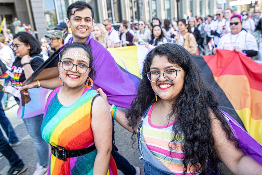 Three people wearing rainbow clothing march in a pride parade, carrying a corner of a very large pride flag.