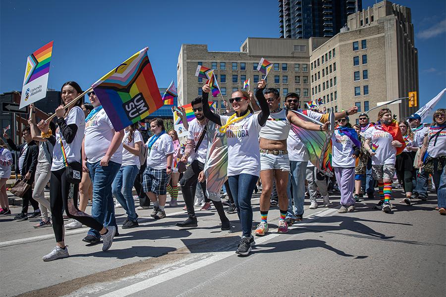 People march in a pride parade in downtown winnipeg. One person carries a pride with the UMSU logo on it.