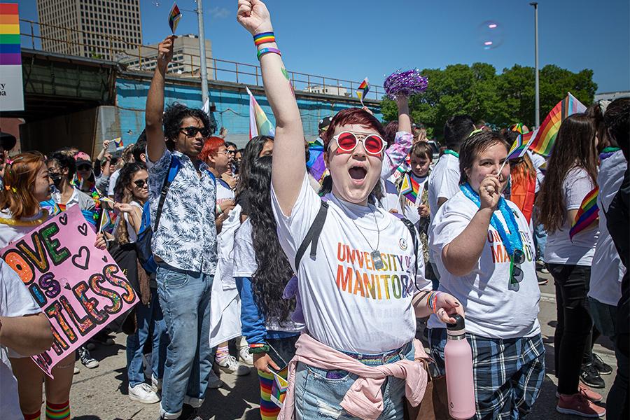 A person wearing a U M rainbow shirt and pink tinted glasses stands with their fist in the air and mouth open in a group of pride marchers.