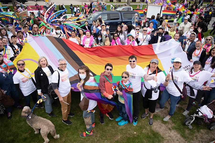 Group of UM staff and students holding a large pride flag. The flag includes a purple circle on yellow background; a white, pink, blue, brown, and black chevron; rainbow horizontal stripes; and the UM logo.