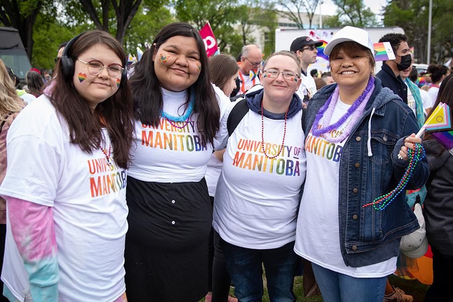 Four smiling people in a larger crowd, wearing U M rainbow t-shirts and heart, butterfly, and rainbow flag temporary tattoos on their cheeks.