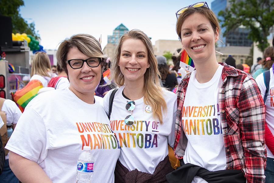 Three smiling people in downtown Winnipeg wearing U M rainbow shirts.