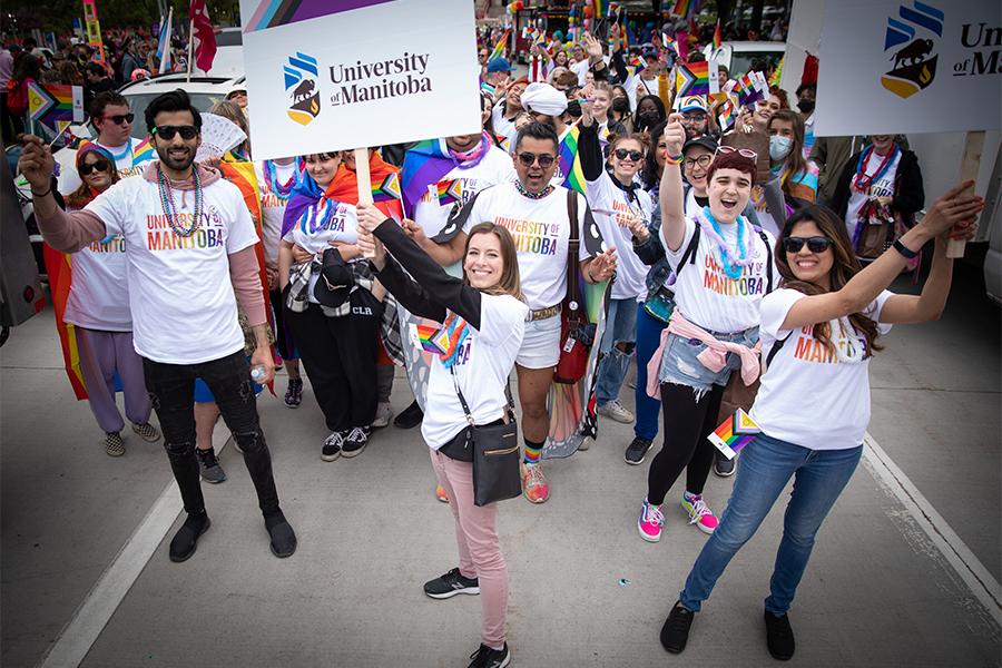 A large group of people wearing U M rainbow shirts and holding pride flags marching in a pride parade.
