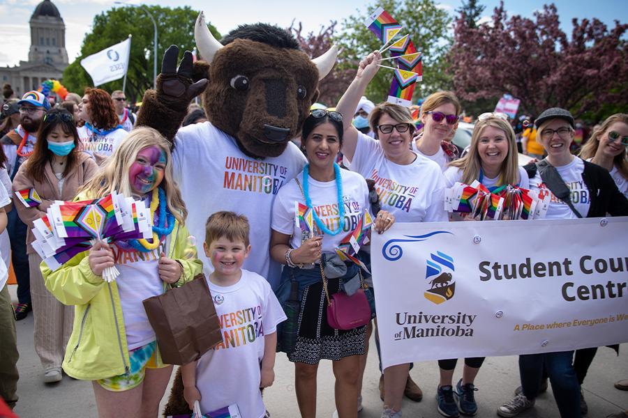 UM mascot Billy the Bison wearing a U M rainbow shirt surrounded by children and adults marching in a pride parade.