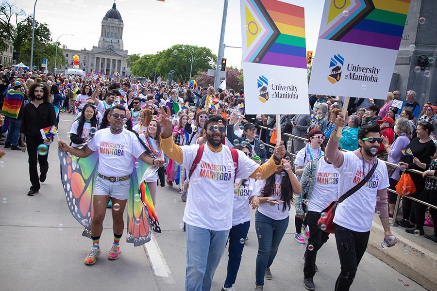 A large group marching in  pride parade. Several are blowing bubbles and carrying U M pride placards. One person is wearing multi-coloured butterfly wings.