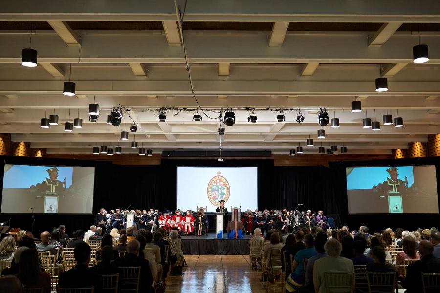 President Benarroch stands at a podium surrounded by numerous seated people on stage, while delivering an address to an audience.