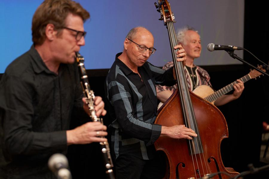 Three men perform on a clarinet, upright bass, and acoustic guitar.