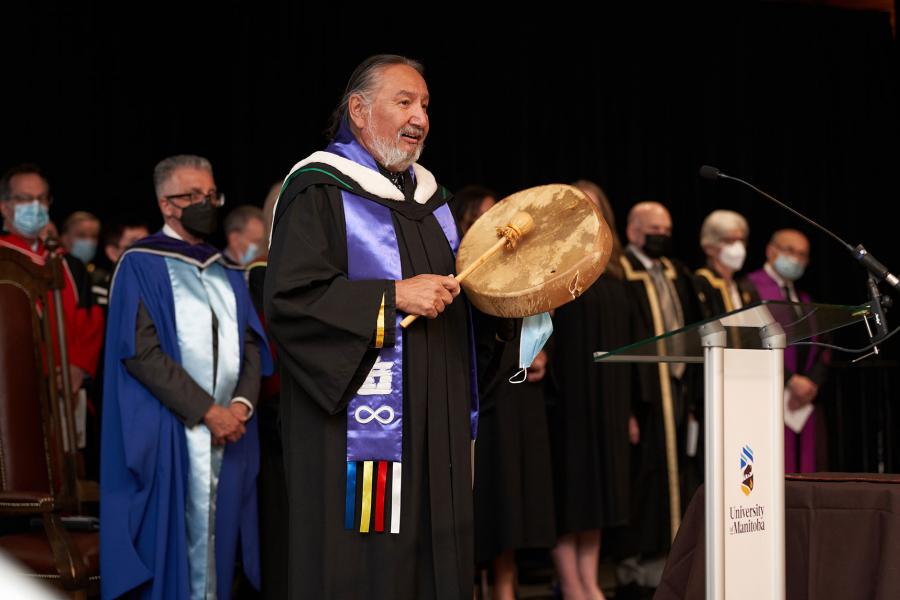 Elder Carl Stone stands wearing an academic gown with Indigenous sash, playing a drum in front of President Benarroch and others.