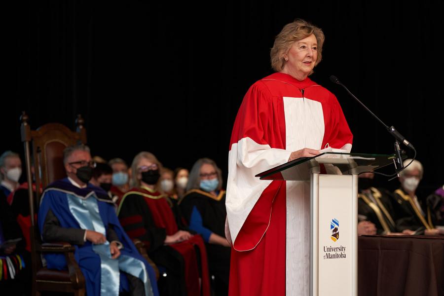 Janice Filmon stands at a podium wearing a red academic gown addressing an audience in front of President Benarroch and others on stage.
