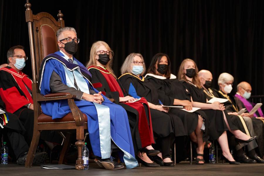 Row of people in academic gowns and covid masks sitting on stage.