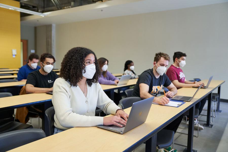 Students wearing KN95 masks and taking notes on laptops and with pencil and paper in a classroom.
