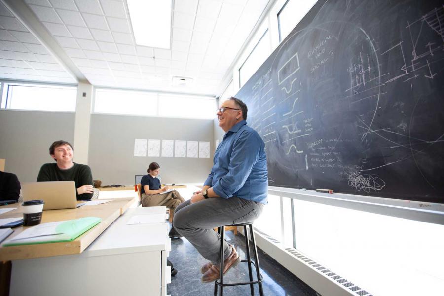 An instructor sits on a stool in front of a blackboard in a brightly lit room with several students.