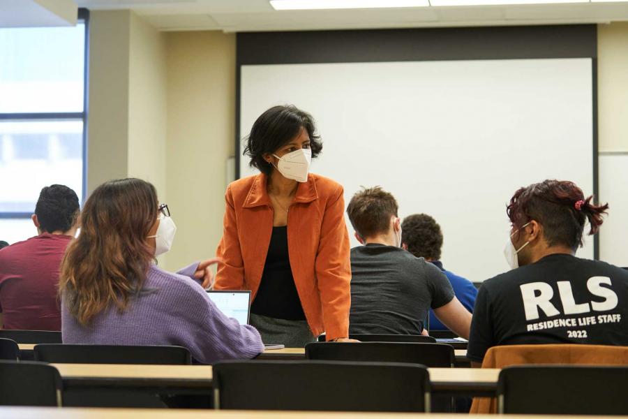 A woman wearing a covid mask stands at a desk in the middle of a lecture hall talking with students.