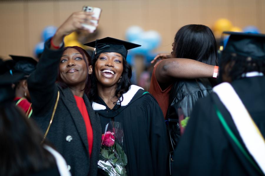 Two women smiling and posing for a selfie. Woman on the right is wearing graduation cap and gown, woman on the left is holding phone.