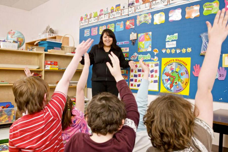 An early years teacher standing at the front of a classroom of kids raising their hands.
