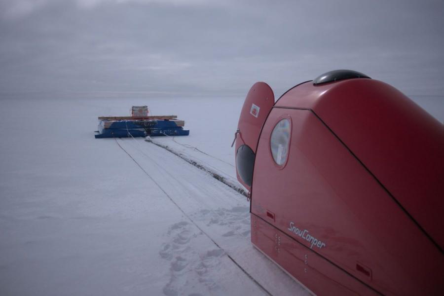 A red snow camper sits on a Canadian Arctic ice field with radar technology equipment in the distance.