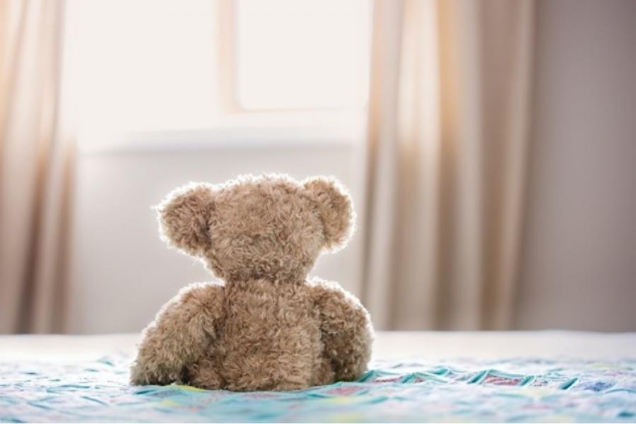 The back of a brown teddy bear placed on a bed in a brightly lit bedroom.
