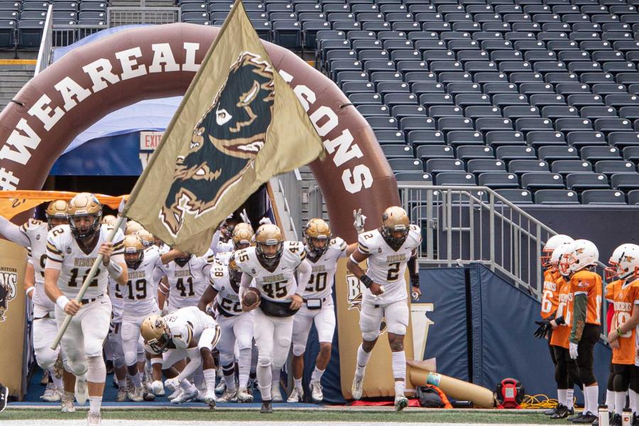 University of Manitoba Bisons football players run onto the field led by a player holding a gold flag with the Bisons logo on it.