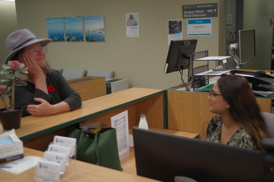 SC Rece[tio desk with receptionist and colleague discussin across desk and hallway to offices and small classroom in the background