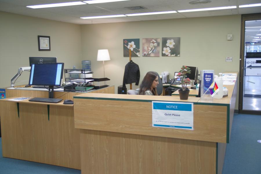 SCC Reception desk view from waiting area with entrance and receptionist in the background and student check-in computer in foreground