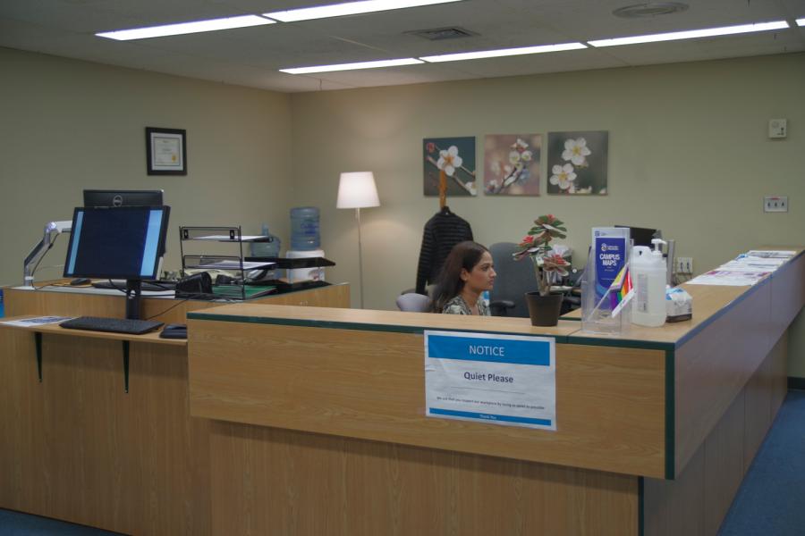 SCC Reception desk view from waiting area with entrance and receptionist in the background and student check-in computer in foreground slighty angled to show hallway to small classroom