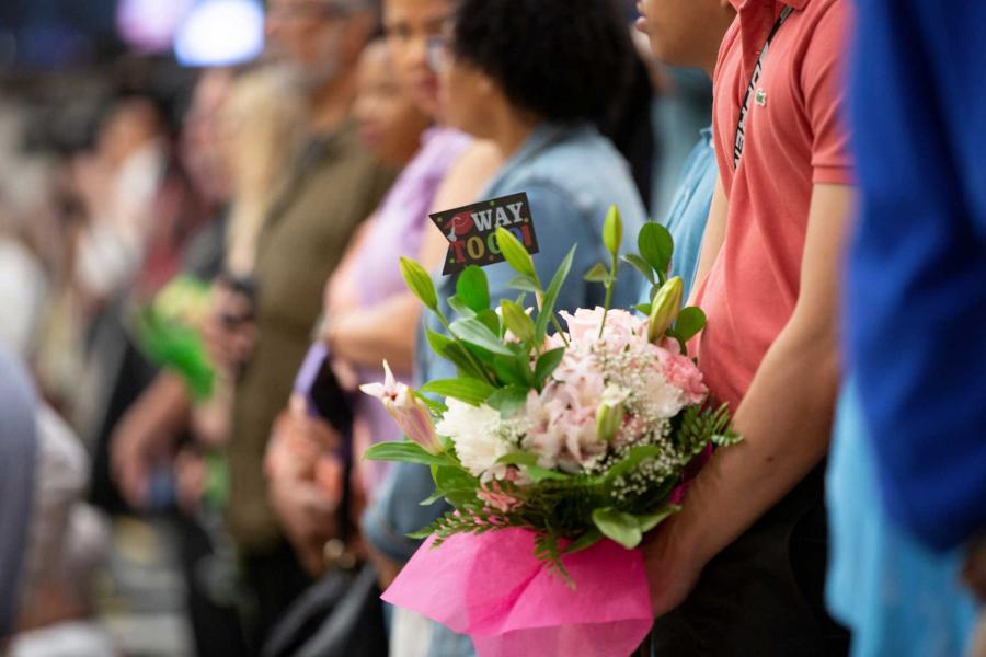 A close-up of a bouquet of pink flowers and a card that says 'way to go!' in someone's arms as they watch a convocation ceremony from the crowd.