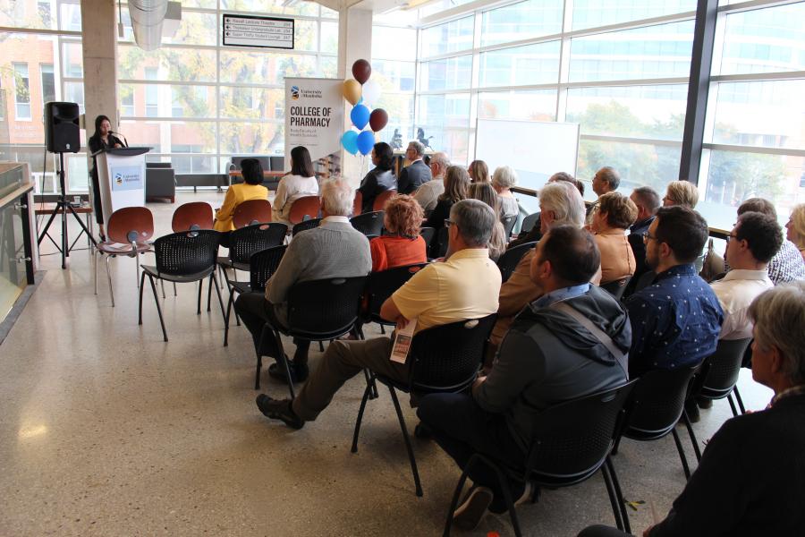 Lady speaking at podicum with guests in chairs facing her in front of College of Pharmacy pop-up banner